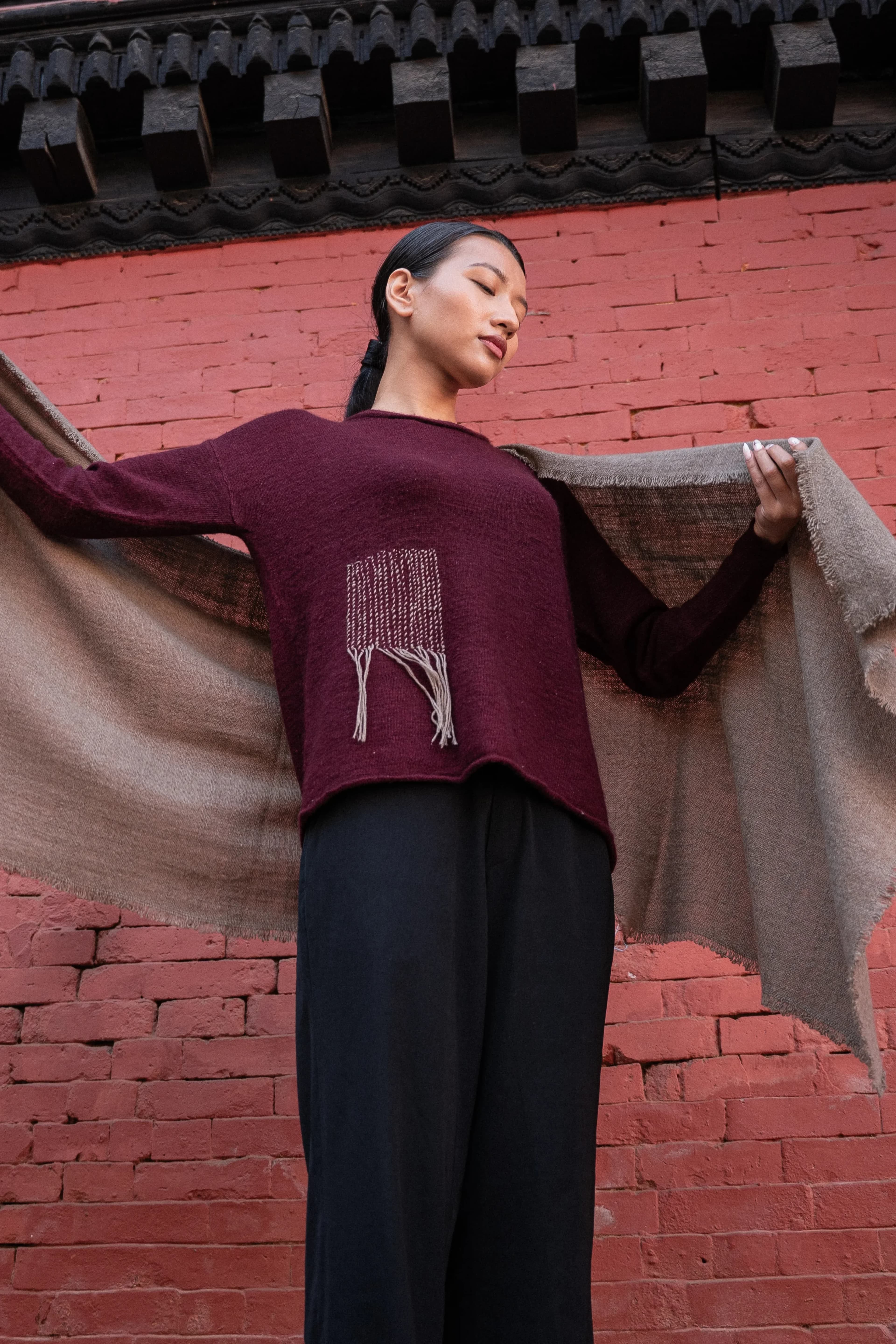 Model in a burgundy knit sweater and black trousers holding a taupe cashmere shawl spread wide against a red brick wall