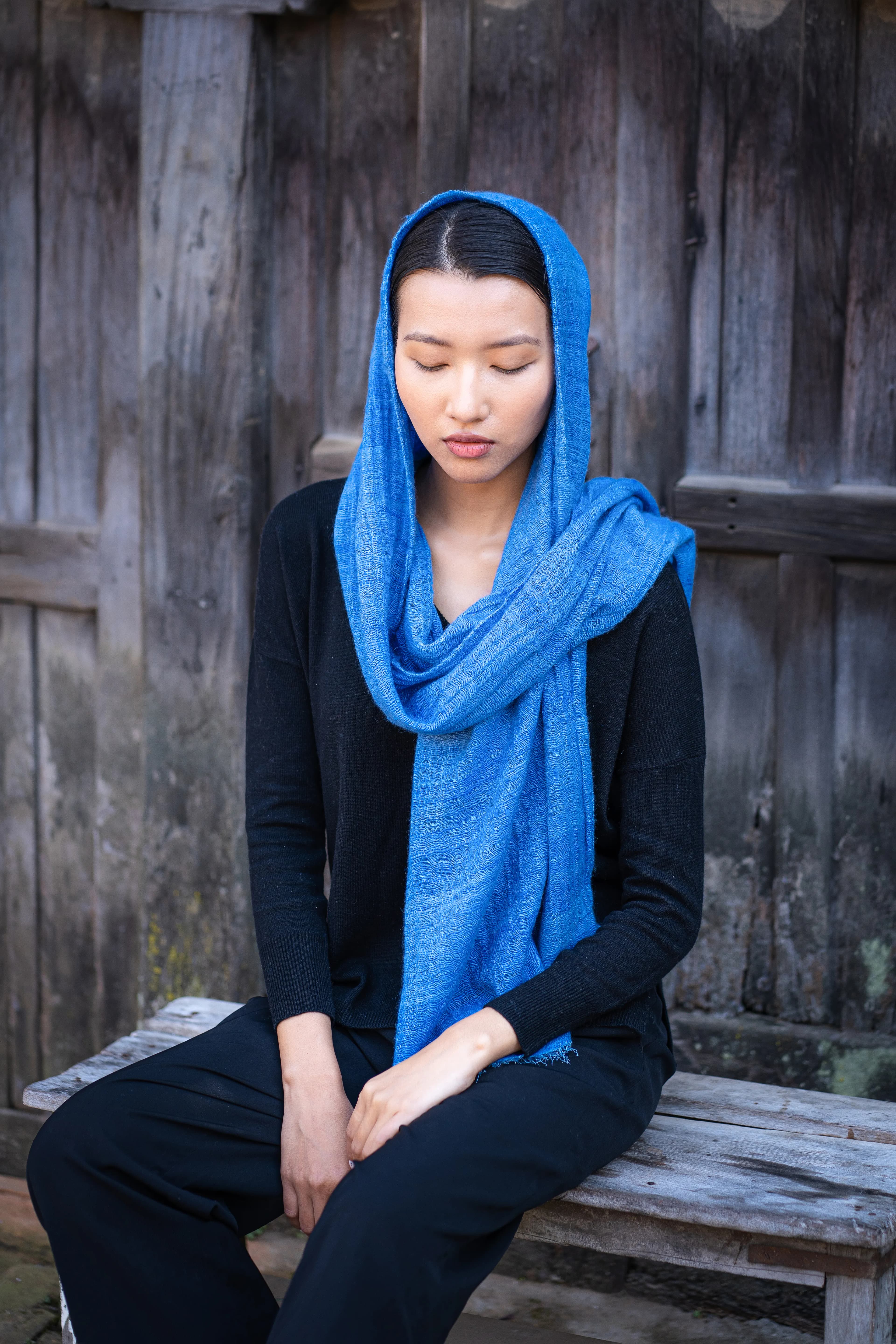 Model seated on a wooden bench with a bright blue cashmere scarf draped over her head and shoulders
