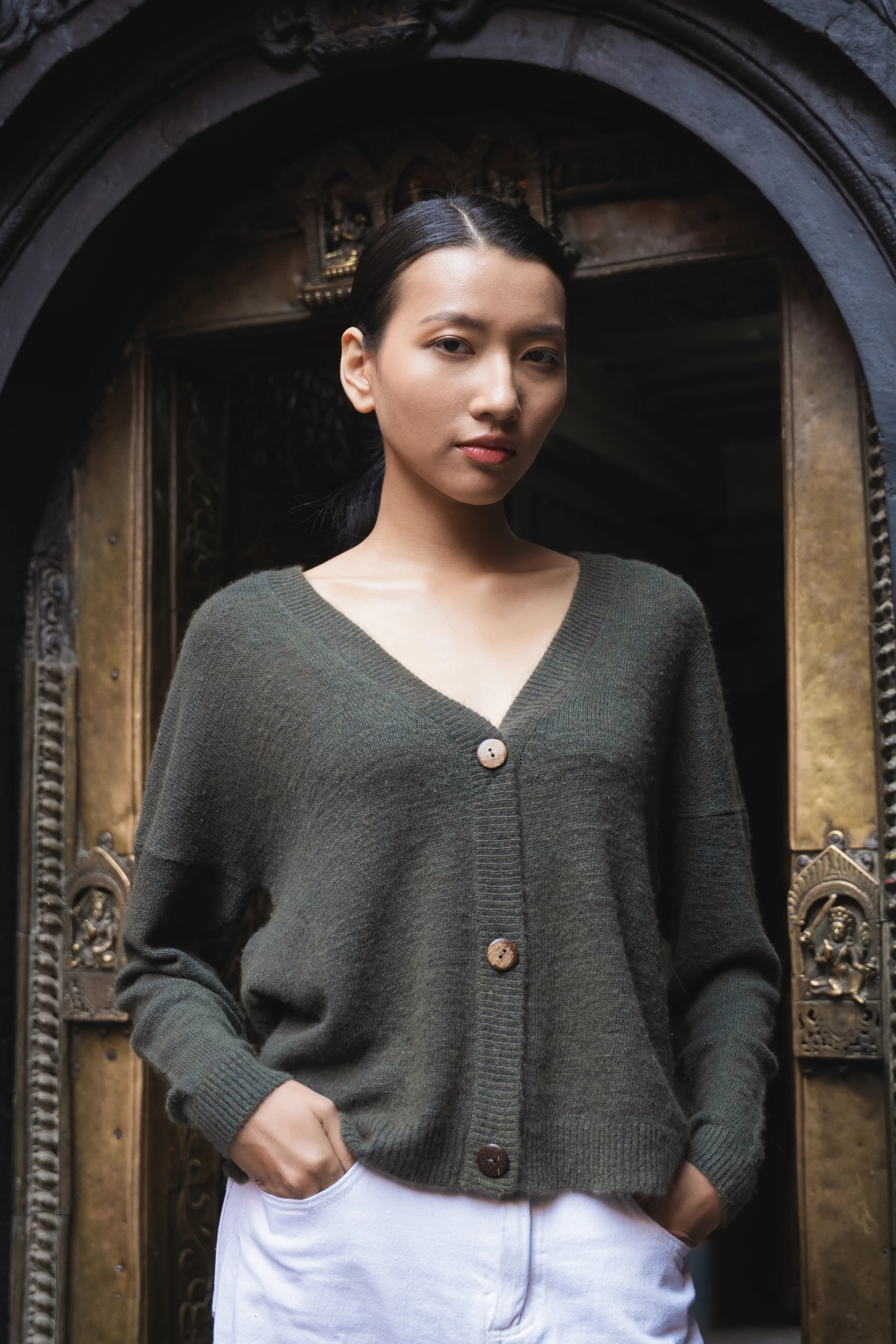 Model in an olive cashmere cardigan with coconut buttons standing in a carved temple doorway