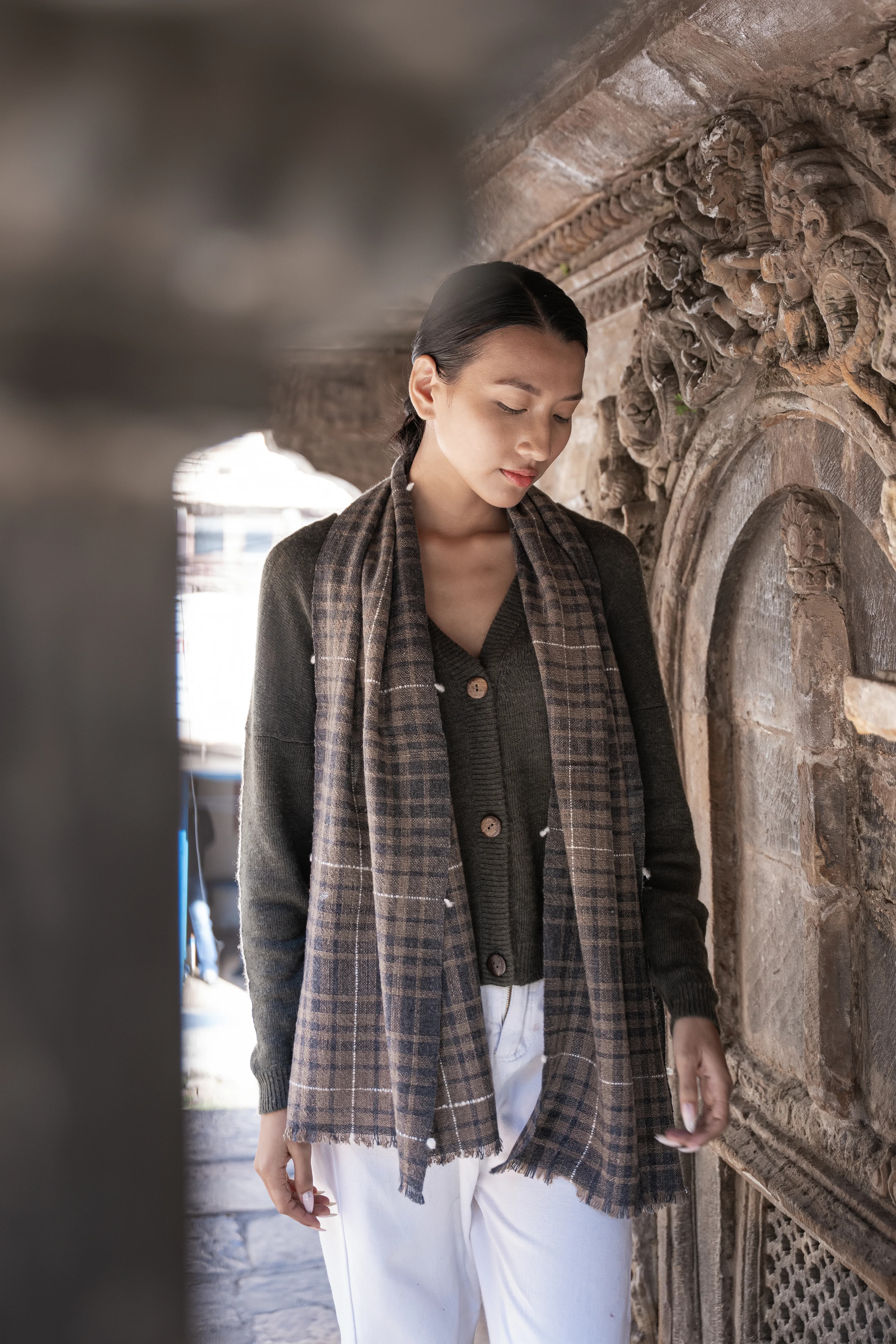 Model wearing a brown and charcoal checked cashmere scarf over an olive cardigan beside a carved stone wall