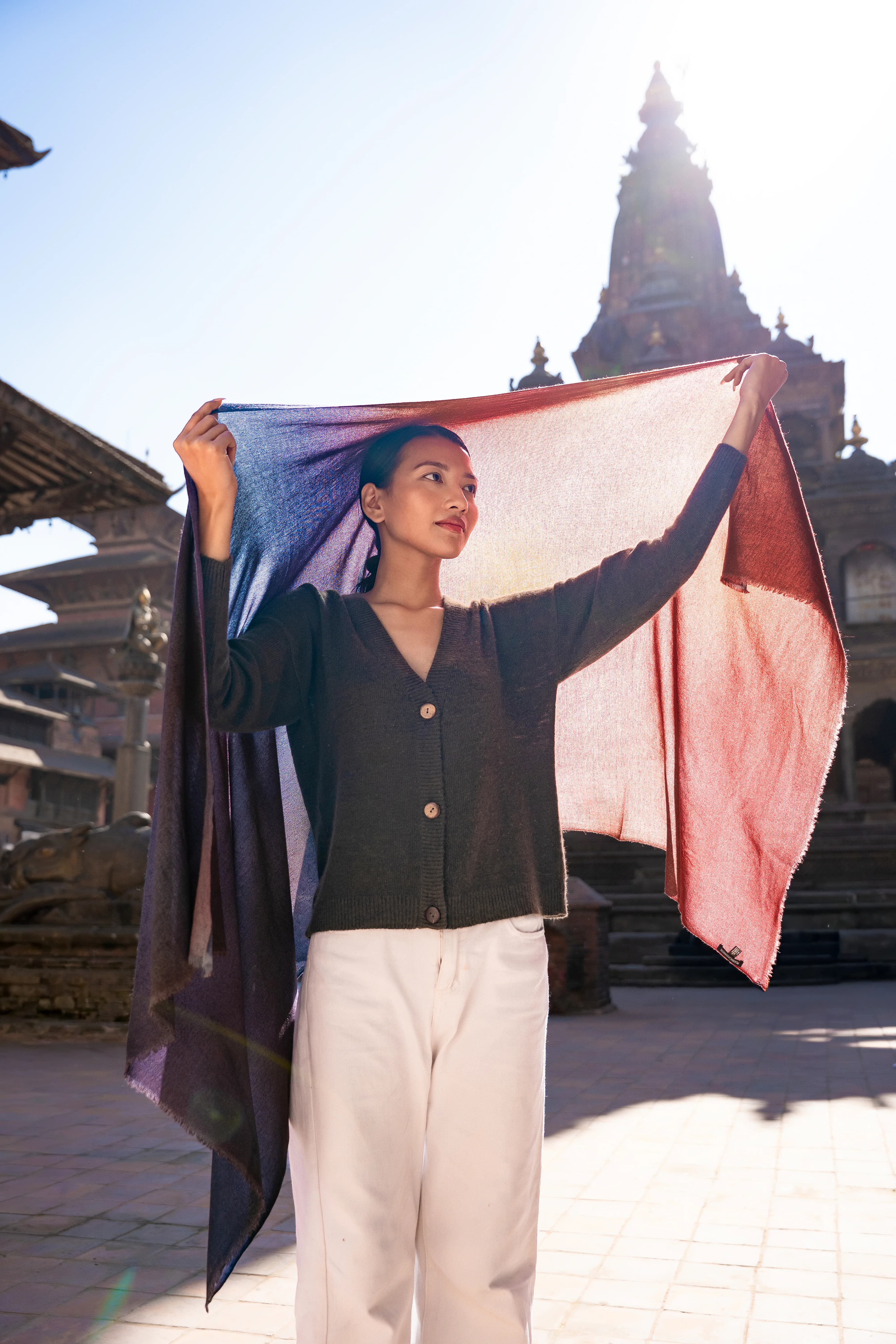 Model holding an ombré blue-to-rust cashmere shawl overhead in a sunlit temple square